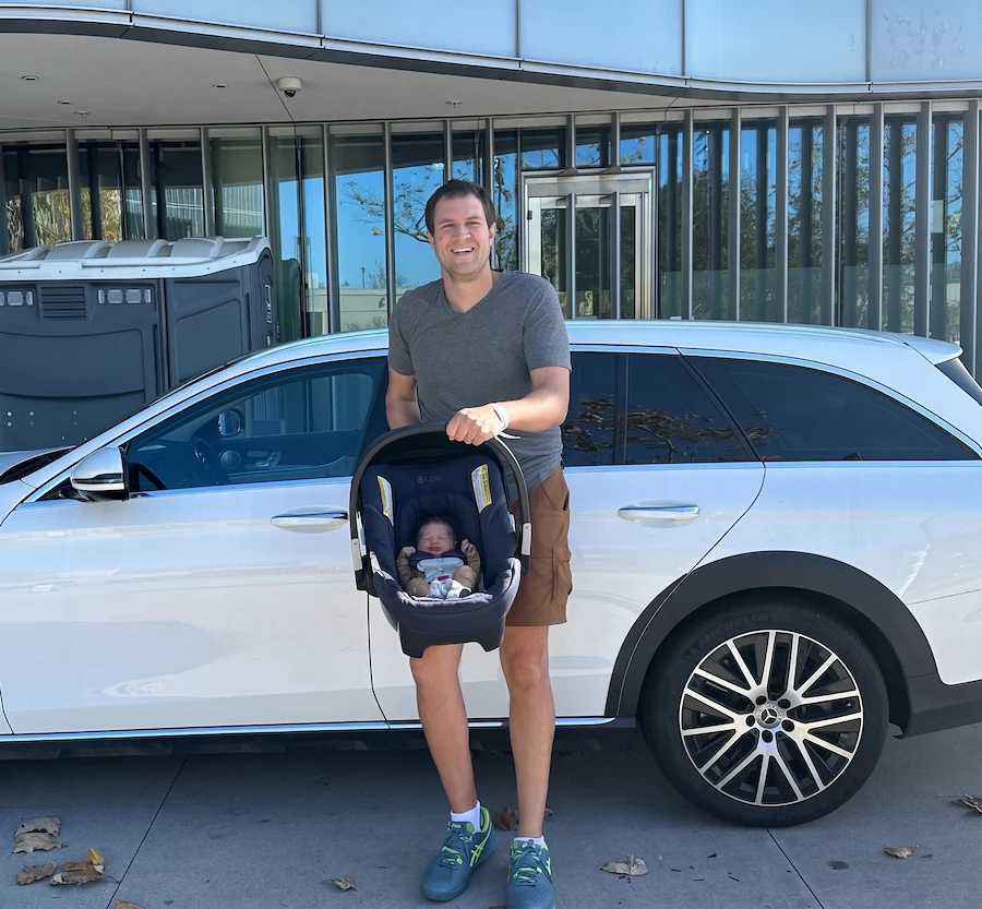 Doug DeMuro holding an infant car seat beside a white Mercedes-Benz wagon, shared on his official Instagram