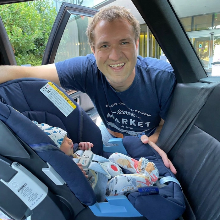 Doug DeMuro smiling beside his newborn son's car seat inside a car, wearing a Sconset Market t-shirt