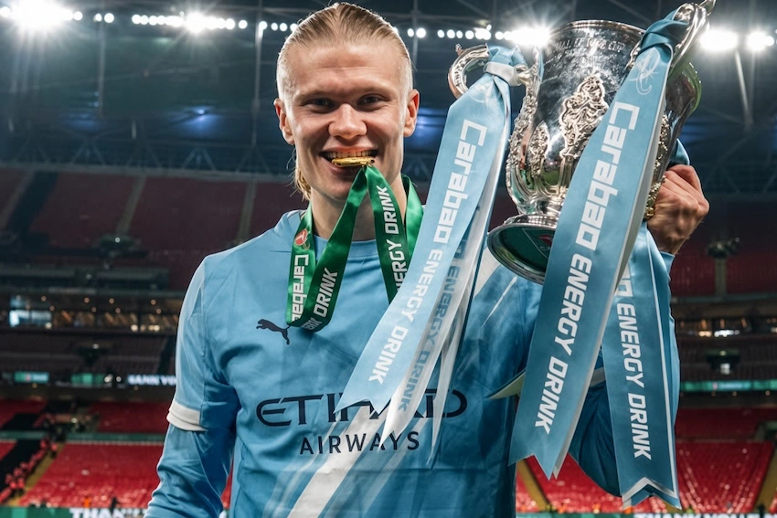 Erling Haaland biting his medal while celebrating with the Carabao Cup in his Manchester City kit