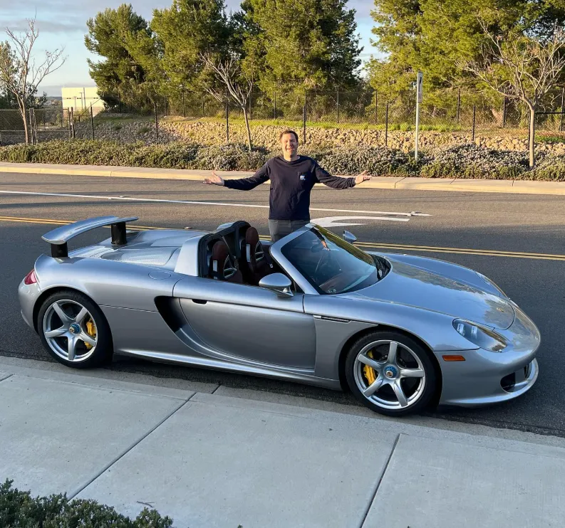 Doug DeMuro standing beside his silver 2004 Porsche Carrera GT with Ascot brown interior, his dream car purchased in February 2023