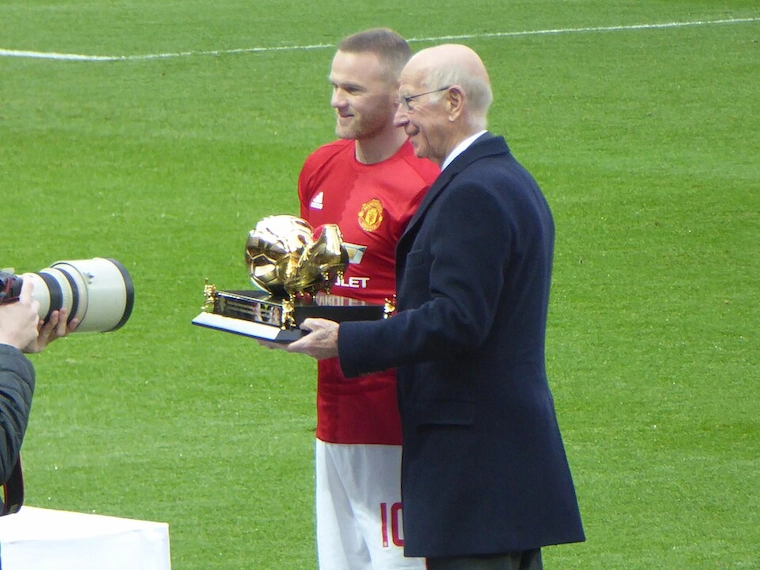 Wayne Rooney in a Manchester United red kit with Sir Bobby Charlton holding a golden trophy at Old Trafford