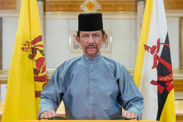Sultan Hassanal Bolkiah at an official address flanked by Brunei's royal and national flags