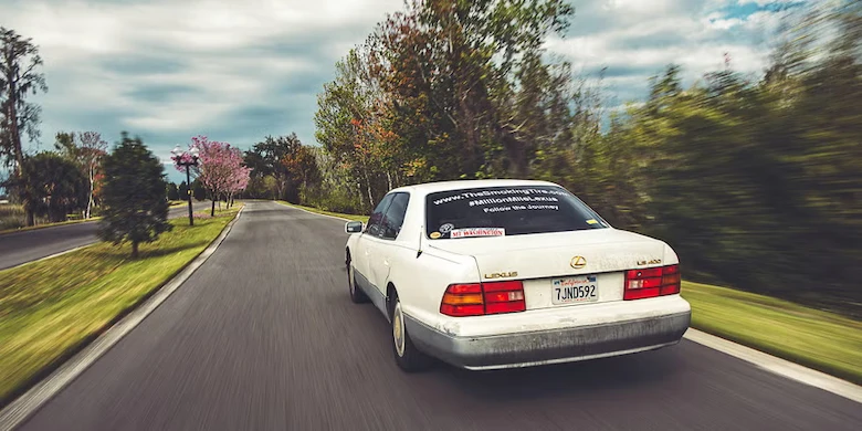 Matt Farah Million Mile Lexus: 1996 LS400 driving down a tree-lined road with project decals on the rear window